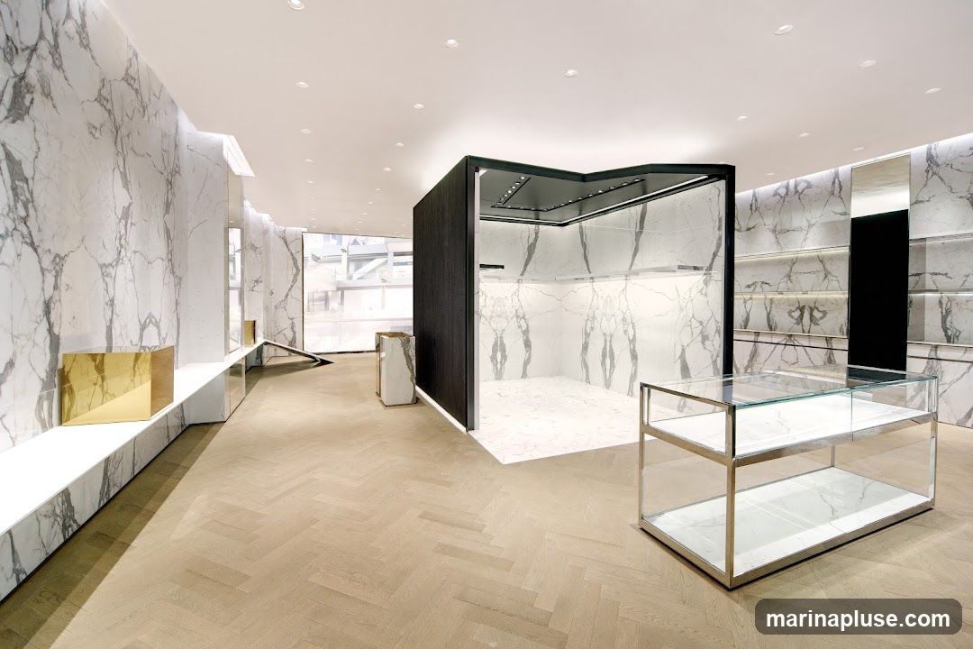 A spacious and minimalist retail interior with marble-patterned walls and light wood herringbone flooring. A central display area features a dark wood frame and marble interior. A glass display case with a metal frame sits in the foreground. Shelving units are visible on the back wall.