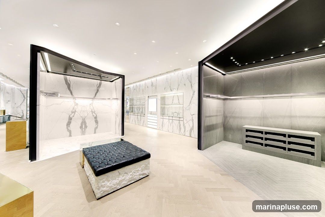A spacious and modern retail interior with marble-patterned walls and light wood flooring. The space features minimalist display units with integrated lighting, a black tufted ottoman, and a grey cabinet with multiple compartments. The overall aesthetic is clean, luxurious, and contemporary, suggesting a high-end fashion or design store.