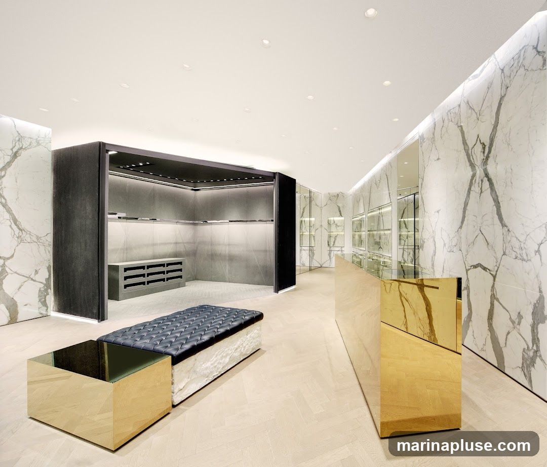 A luxurious and minimalist retail store interior featuring marble-patterned walls, a golden reception desk, and a dark upholstered bench with a mirrored coffee table. The display areas are empty, with shelves and racks visible, suggesting a high-end fashion or accessory boutique. The lighting is bright and modern, with recessed ceiling lights and illuminated wall panels.