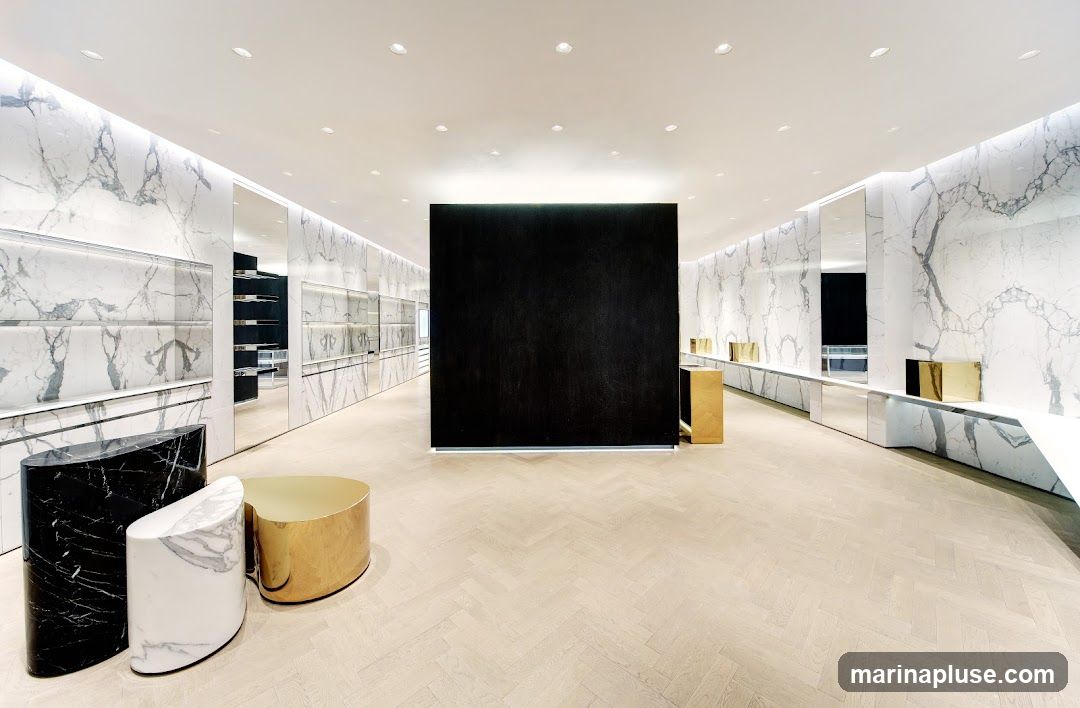 A spacious and minimalist retail store interior with marble-patterned walls, white ceilings with recessed lighting, and light wood herringbone flooring. In the foreground, there are three sculptural display stands: one black and white marble, one white marble, and one gold. A large black rectangular structure is centrally located. The walls feature shelves and mirrored surfaces, with gold decorative items displayed on a long white counter.