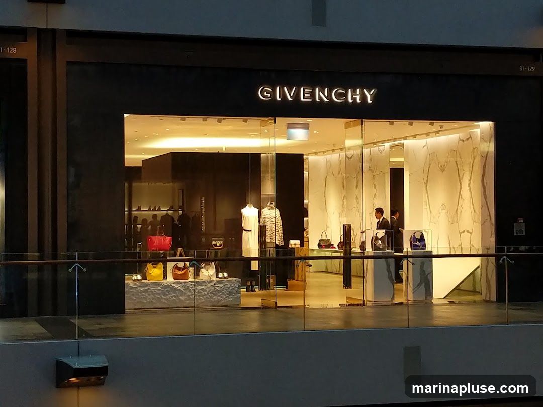 The exterior of a Givenchy store with its name prominently displayed above the entrance. The store features a modern design with large glass windows showcasing handbags and clothing displays. Two staff members are visible inside the store.