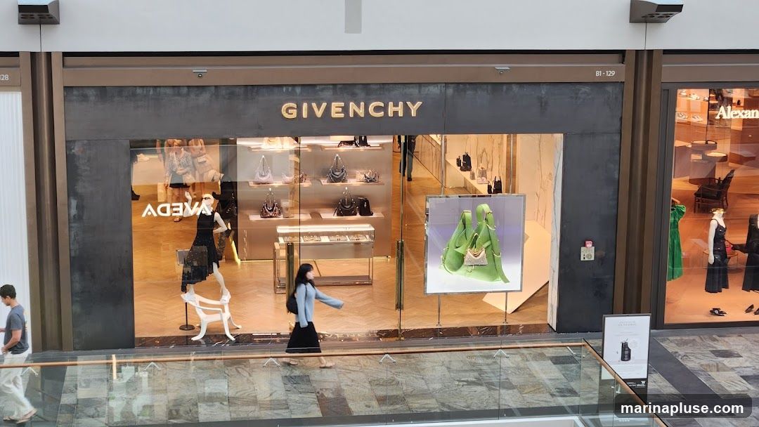 The exterior of a Givenchy store with the brand name prominently displayed above the entrance. The storefront features large glass windows showcasing handbags and accessories on display shelves and mannequins. A large display of a green handbag is visible to the right. People are seen walking past the store and inside.