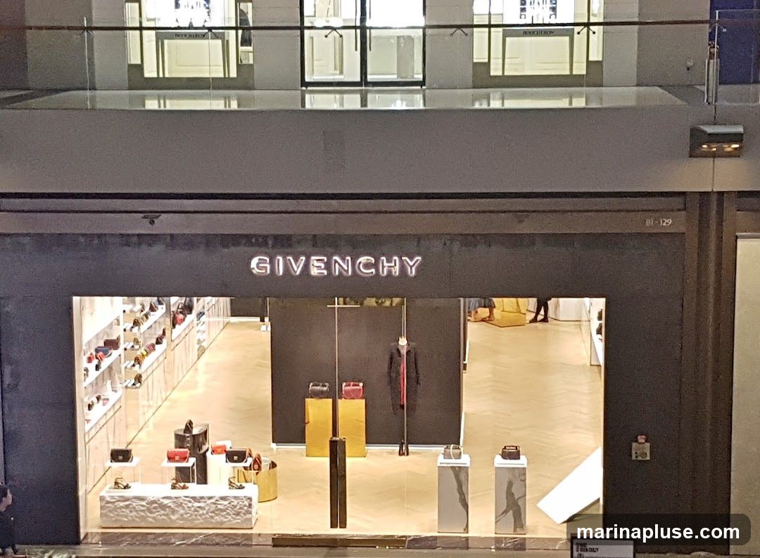 A wide shot of the Givenchy store entrance. The store name is displayed in large, illuminated letters above the entrance. The storefront features a large display window showcasing handbags and shoes on shelves and pedestals. Inside, a mannequin is dressed in a dark coat with a red scarf. The interior has a polished wooden floor.