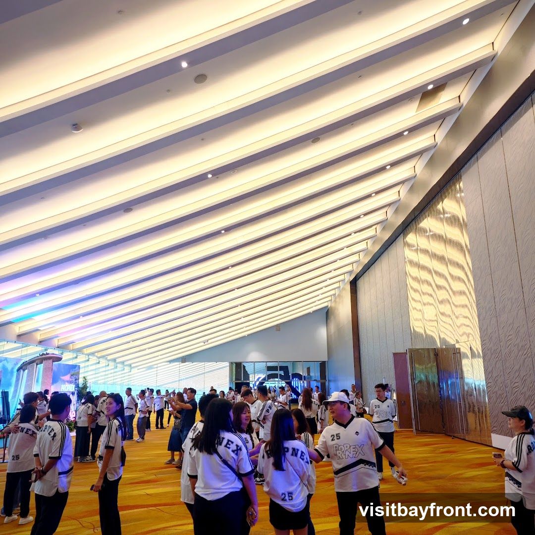 A large group of people are gathered in a spacious hall with a distinctive sloped ceiling featuring parallel lighting elements. Many individuals are wearing matching white t-shirts with black accents and lettering, suggesting a team or event affiliation. The hall has a warm-toned carpeted floor and textured walls. In the background, there are some displays and entrances, with colorful lighting visible on the left side. The overall atmosphere appears to be that of a conference, event, or gathering.
