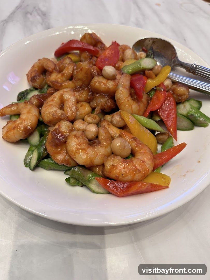 A white plate filled with a stir-fry dish featuring shrimp, asparagus, red and yellow bell peppers, and peanuts in a glossy sauce. A spoon and fork rest on the side of the plate. The dish is presented on a marble-patterned surface.