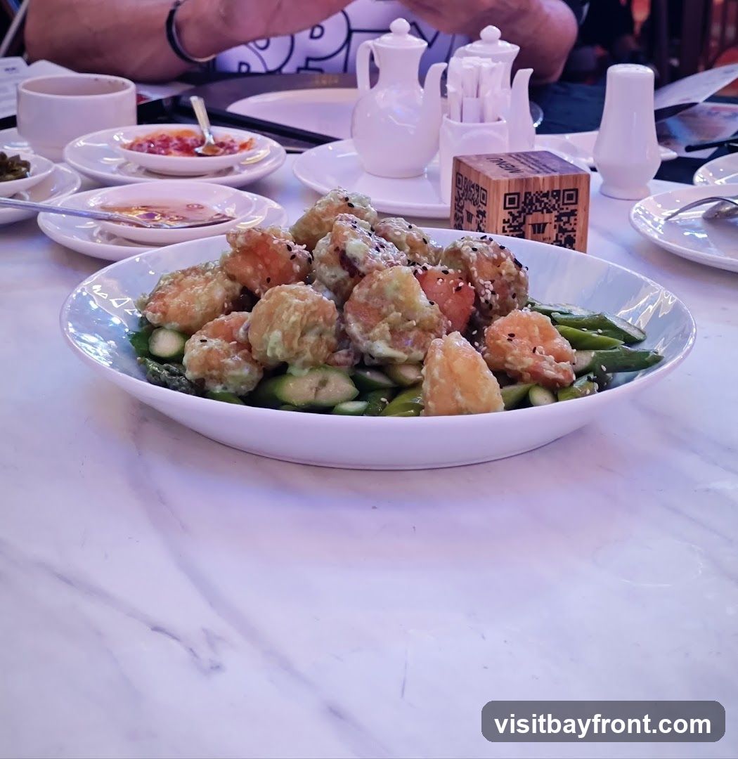 A close-up shot of a dish featuring fried shrimp and asparagus, drizzled with a creamy sauce and sprinkled with sesame seeds. The dish is served in a white oval bowl on a marble-patterned table. In the background, there are various tableware items, including small dipping bowls, teapots, and a salt shaker. A wooden cube with a QR code and the word "MENU" is also visible on the table.
