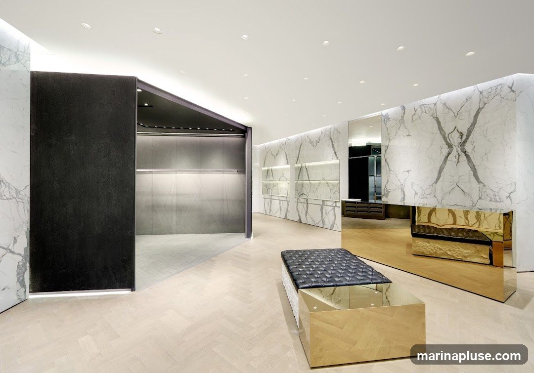 A spacious and minimalist retail interior with marble-patterned walls, light wood herringbone flooring, and sleek display areas. A black upholstered bench with a mirrored base sits in the foreground, reflecting the polished gold counter and the surrounding decor. The space appears to be a high-end boutique or showroom, with empty clothing racks visible in one section.