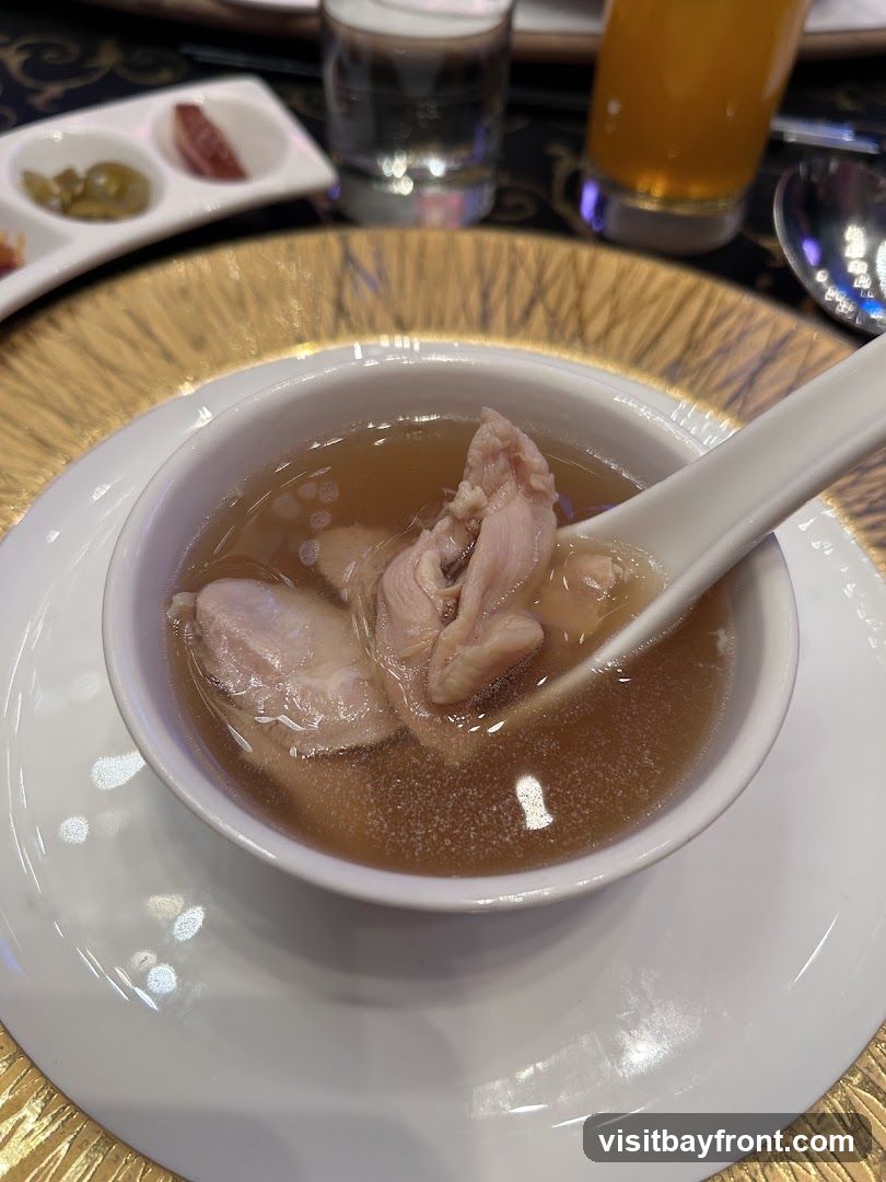 A close-up shot of a bowl of chicken soup with pieces of chicken visible. The soup is in a white bowl, which is placed on a white plate with a gold rim. In the background, there are small dishes with appetizers, a glass of water, and a glass of orange juice.