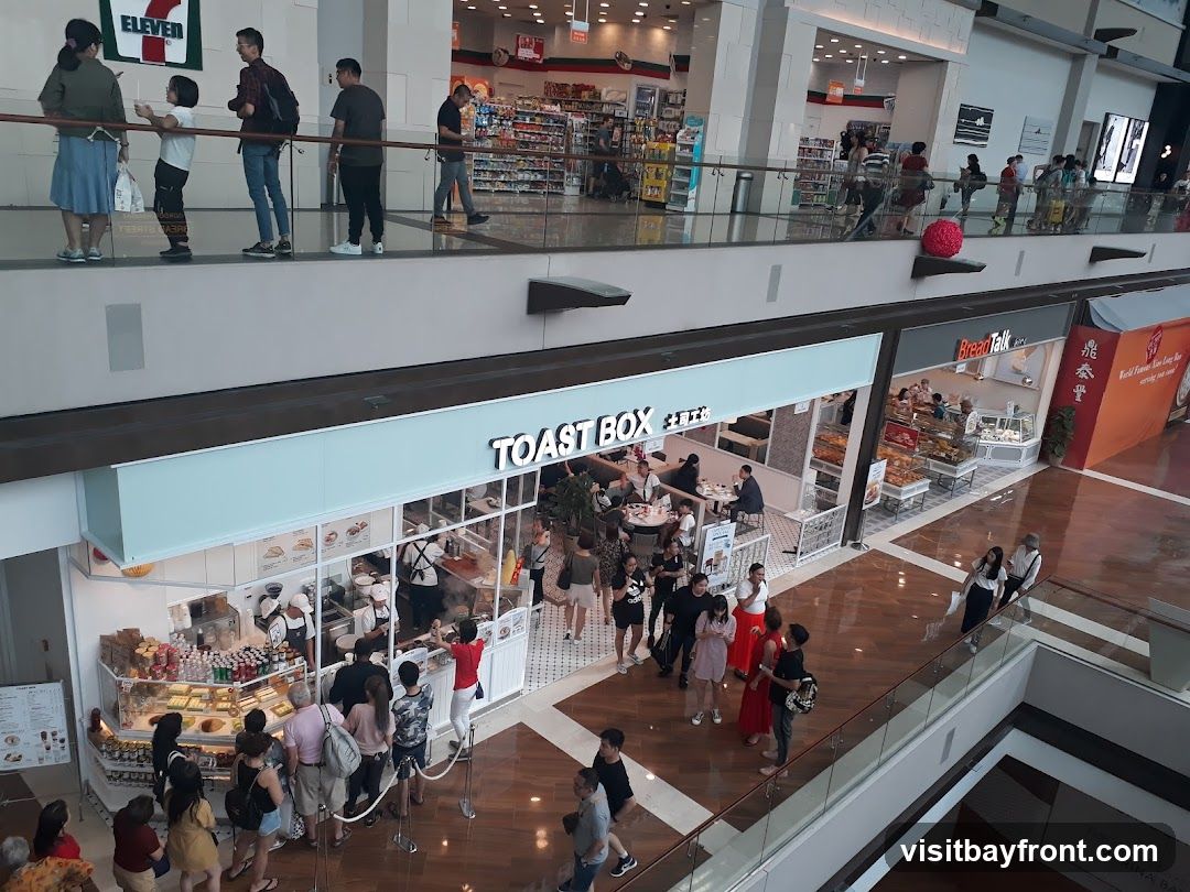 An overhead view of a busy mall food court. In the foreground, a shop called TOAST BOX is visible with customers queuing and dining inside. To the right, a shop labeled BreadTalk is also seen with display cases of baked goods. Further in the background, a 7-Eleven store is identifiable by its prominent signage. People are seen walking and browsing on different levels of the mall.