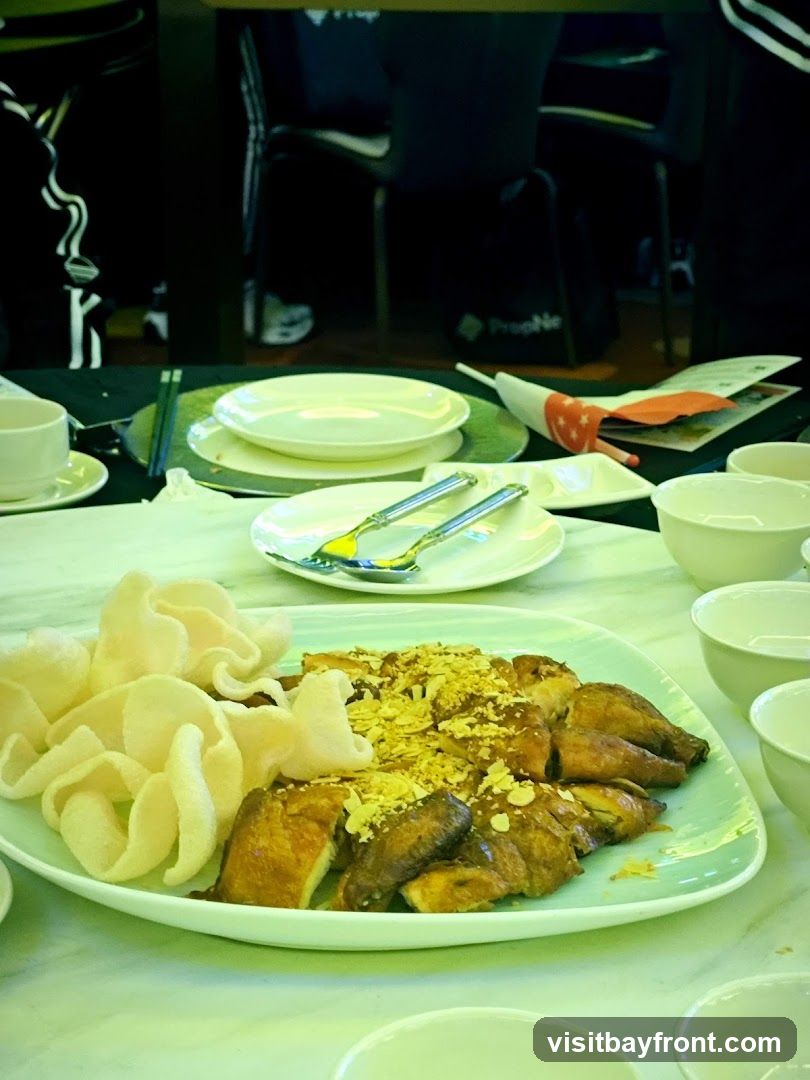 A platter of sliced roasted chicken topped with chopped nuts and served with a side of prawn crackers. The dish is presented on a light green rectangular plate, set on a table with white bowls, plates, and chopsticks. In the background, blurred figures are seated at tables, suggesting a restaurant setting. A small Singaporean flag is visible on the table.
