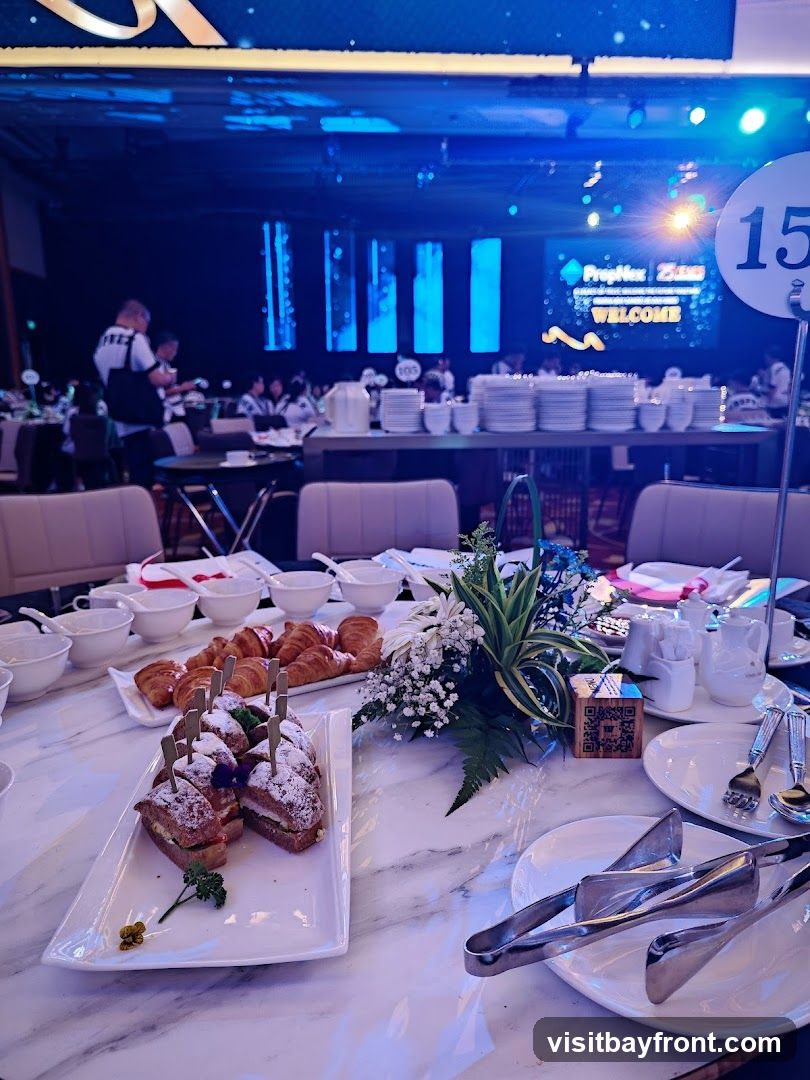 A table set for an event with a spread of pastries and sandwiches. In the background, a stage with a screen displaying "PropNex 25th Anniversary WELCOME" and people attending the event.