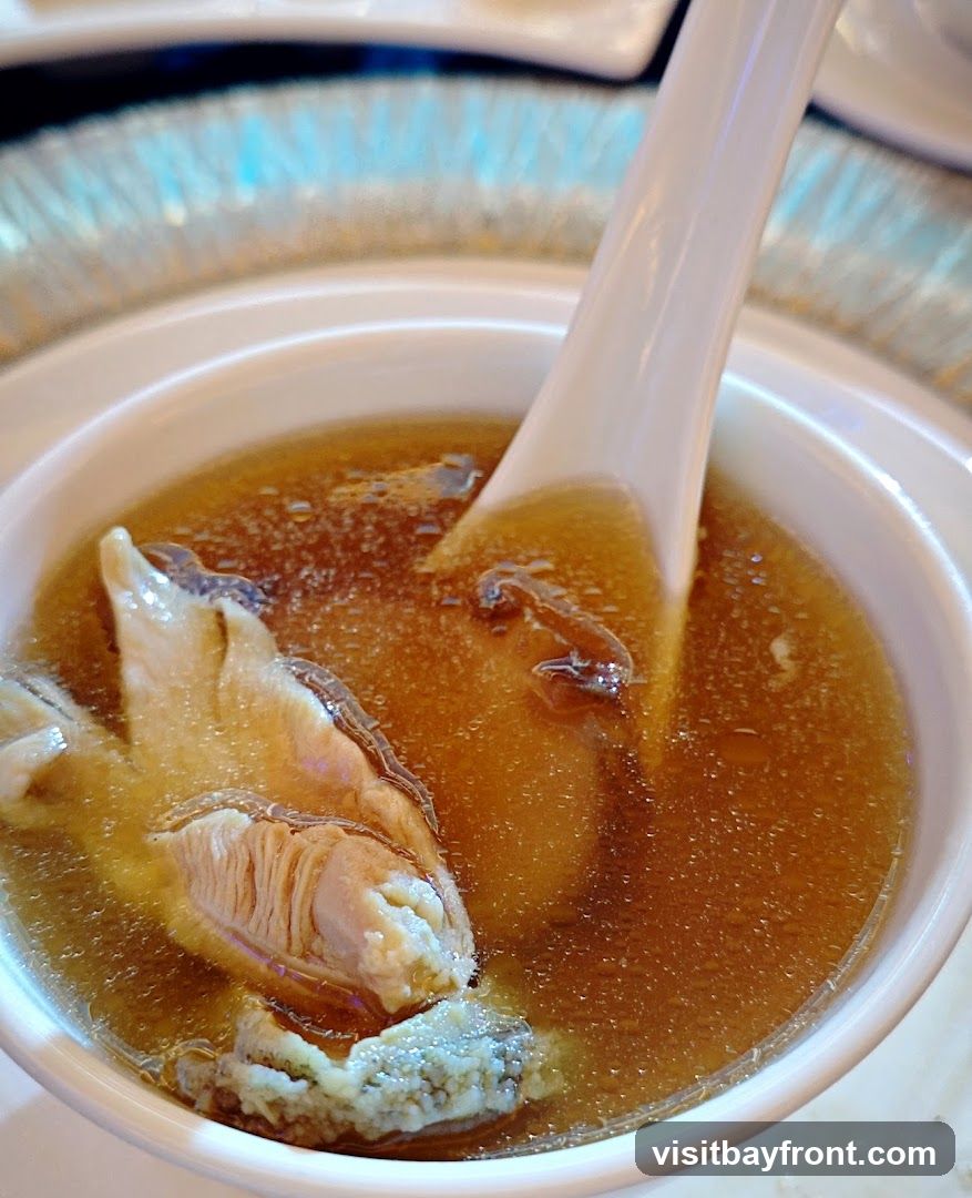 A close-up shot of a white bowl filled with a rich, amber-colored soup. A white ceramic spoon is partially submerged in the liquid, and a piece of meat, possibly chicken or fish, with visible muscle fibers, is prominent in the foreground. The soup appears to have small, dark pieces floating within it. The background is blurred, showing hints of a decorative, possibly metallic, placemat or table setting with blue accents.