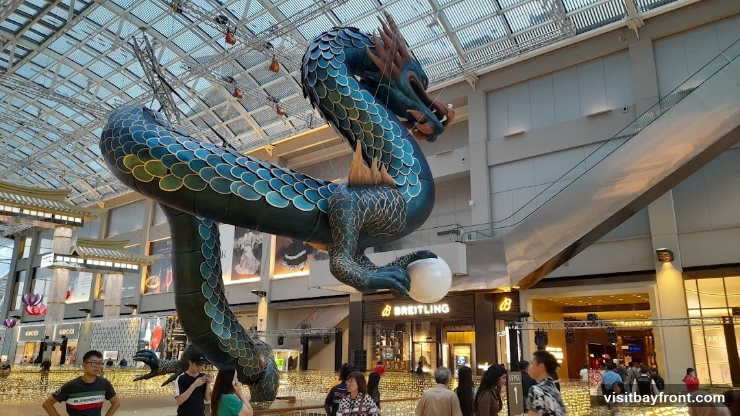 A large, ornate dragon sculpture hangs suspended from the ceiling of a modern shopping mall. The dragon is predominantly teal and dark blue with gold accents, featuring intricate scales and sharp claws. It is coiled around a large white sphere. Below the dragon, escalators and various shop fronts are visible, including a Breitling store. People are seen walking around the mall, and decorative lights are strung throughout the lower levels. The mall has a high, glass-paneled ceiling that allows natural light to filter in.