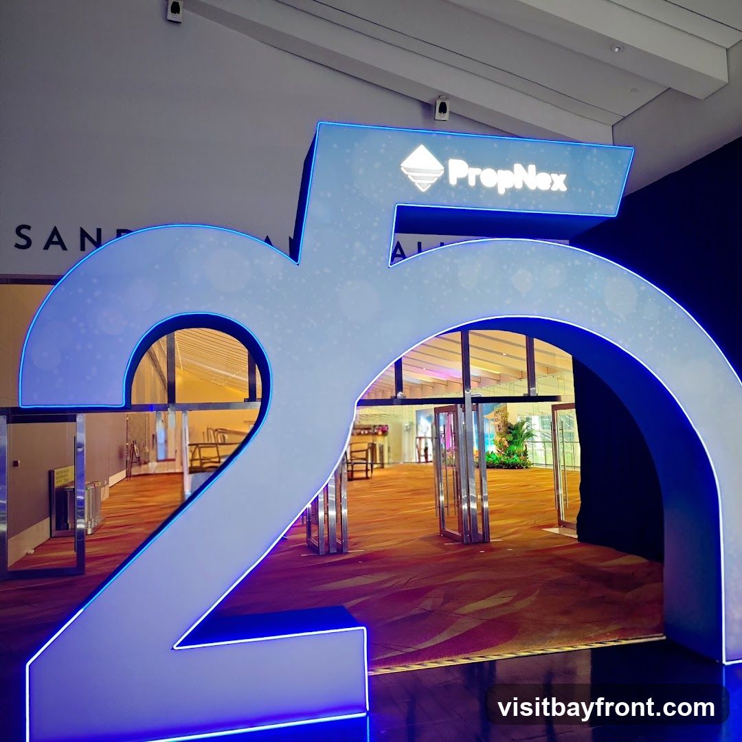A large, illuminated archway shaped like the number 25, with the PropNex logo and name displayed on the top bar. The archway is blue and white with neon blue trim. Beyond the archway, an indoor event space with glass doors, seating, and tropical plants is visible. The floor has a patterned carpet.