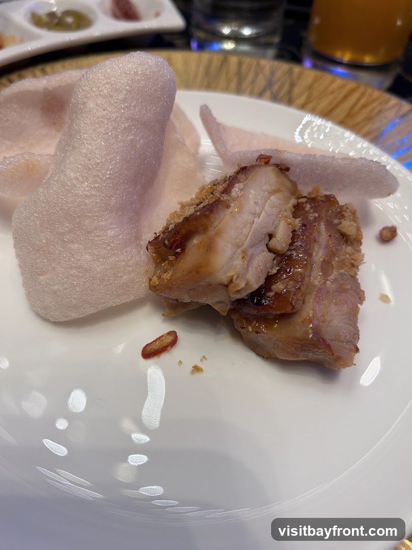 A close-up shot of a plate of food featuring several pieces of glistening, glazed pork belly served alongside light pink, puffed prawn crackers. A few small, sliced red chilies and crumbs are scattered on the white plate. In the blurred background, there are small dishes with dips and a glass of orange liquid.