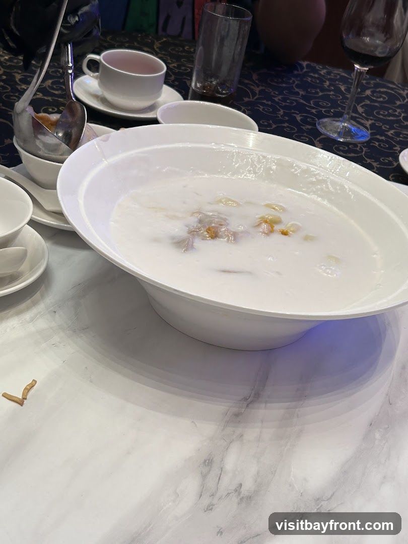 A white bowl filled with a creamy dessert, possibly a sweet soup or pudding, is placed on a marble-patterned table. The dessert appears to have some solid pieces within it. A ladle rests in a smaller white bowl to the left of the main bowl. In the background, there are white teacups, a glass, and a wine glass, suggesting a dining setting.