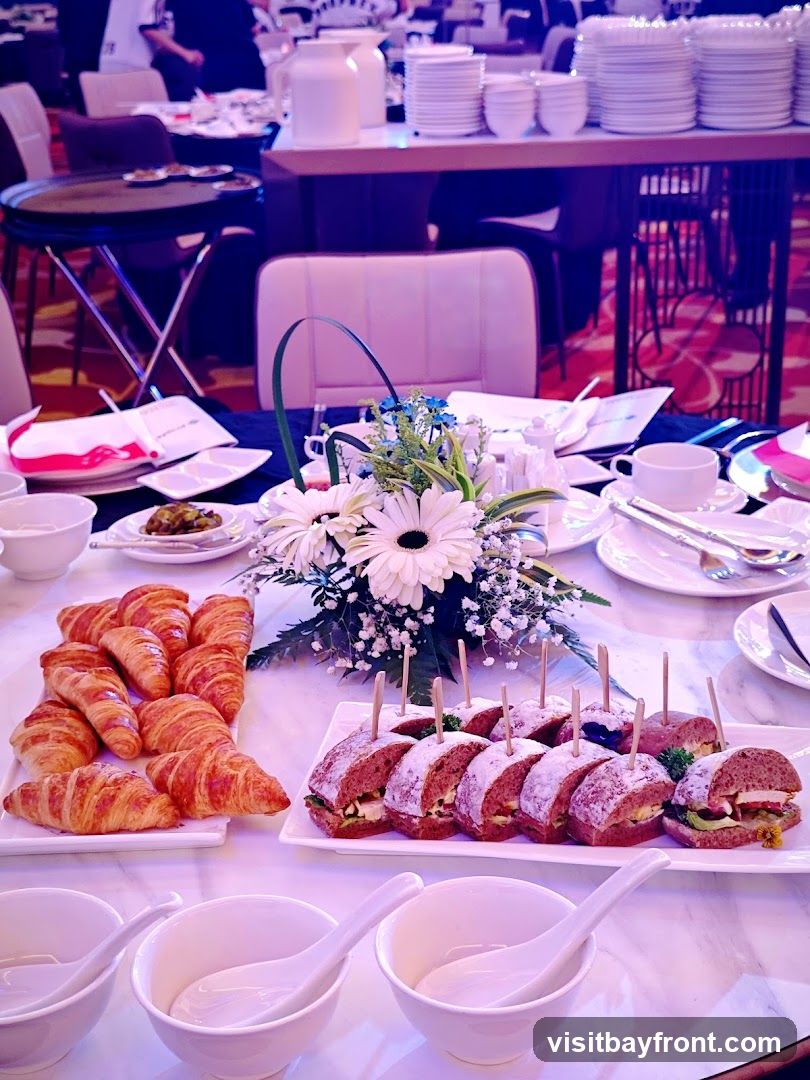 A close-up view of a buffet table set for an event. In the foreground, there are white bowls with spoons. To the left, a platter holds numerous golden-brown croissants. To the right, a rectangular white platter displays an assortment of small sandwiches, each topped with a wooden skewer. A floral centerpiece with white gerbera daisies and delicate white flowers adds a touch of elegance. In the background, stacked plates and pitchers are visible on a shelf, and blurred figures of people can be seen seated at tables in a dining area. The lighting casts a pinkish-purple hue over the scene.
