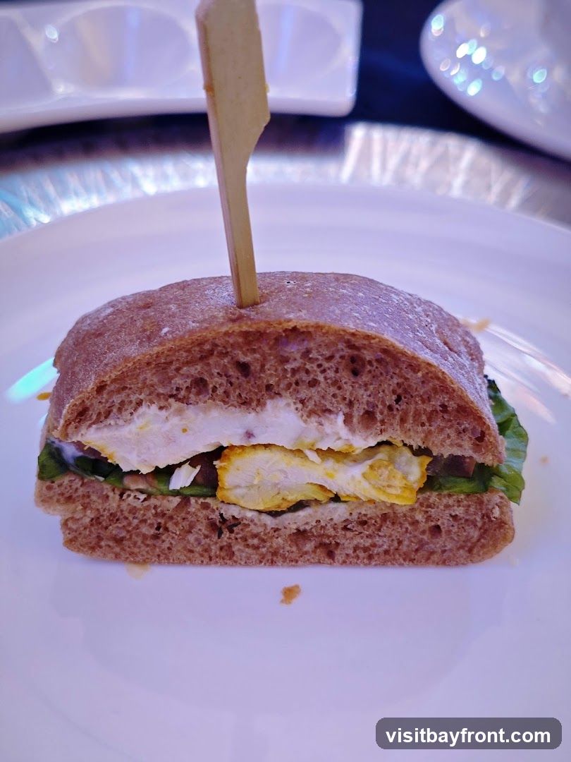 A close-up shot of a chicken sandwich on a white plate. The sandwich is made with dark bread and filled with lettuce, chicken pieces, and a white sauce. A wooden skewer holds the sandwich together.