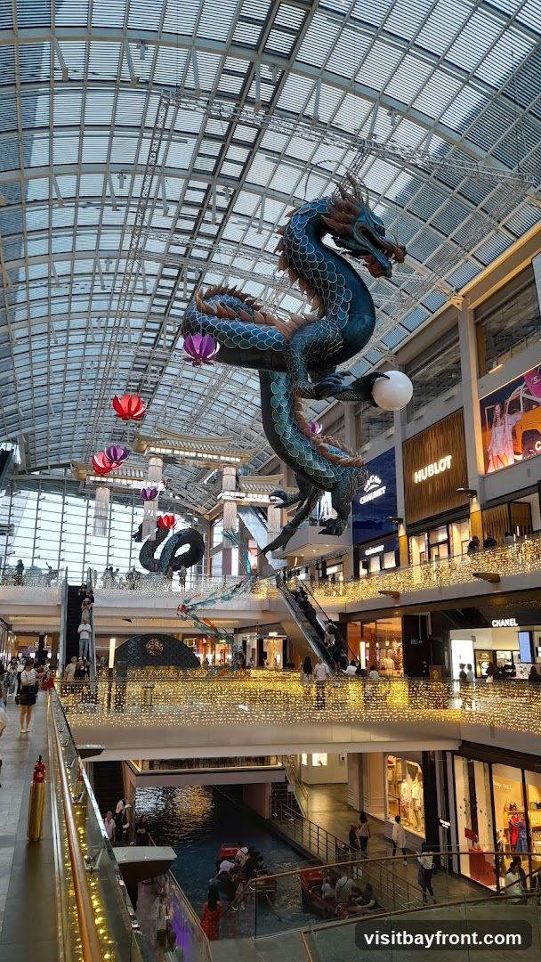 A large, multi-level shopping mall interior is decorated with a large, blue dragon sculpture suspended from the ceiling. Colorful lanterns in the shape of flowers are also hanging. The mall features escalators, shops with illuminated signage (e.g., HUBLOT, CHANEL), and a canal with gondolas on the lower level. Many shoppers are visible on the different levels. The ceiling is a glass and metal structure.