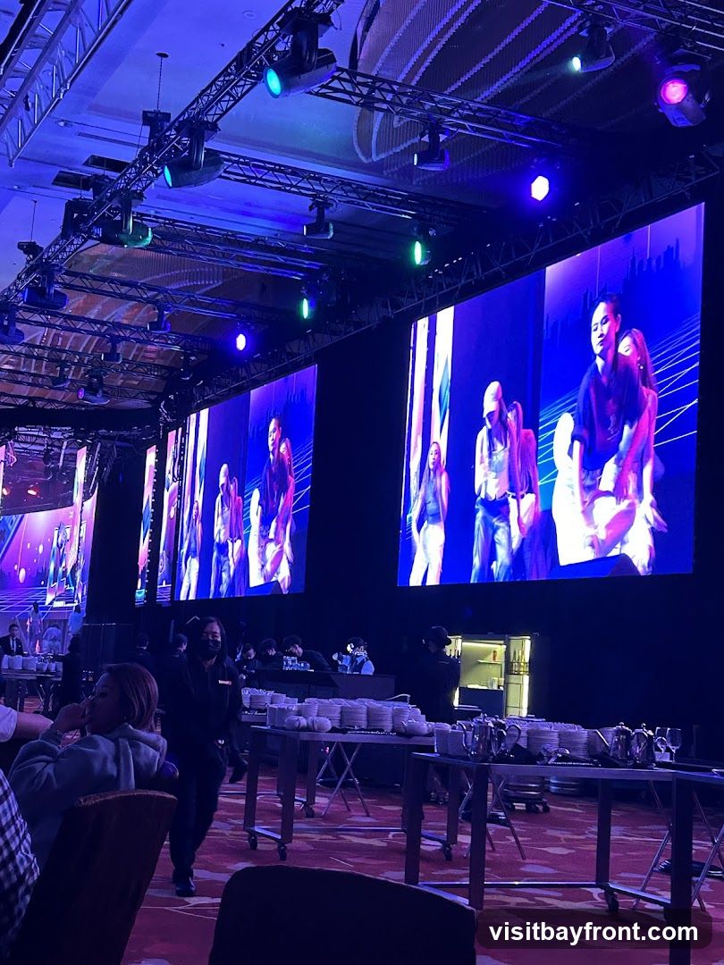 A large indoor event space with multiple large screens displaying images of people. The screens are illuminated by stage lighting. In the foreground, tables are set up with stacks of plates and cutlery, suggesting a catering service. Several people are visible, some seated and some standing, including staff members wearing masks. The overall ambiance is that of a formal gathering or performance.