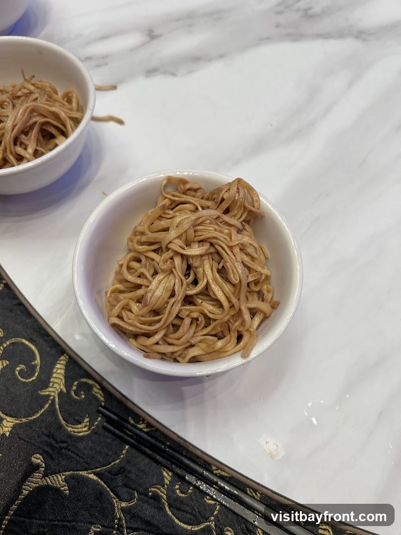 A close-up shot of a white bowl filled with cooked noodles. The noodles appear to be thin and have a light brown sauce. Another bowl of noodles is partially visible in the upper left corner. A black placemat with a gold ornate pattern and chopsticks are in the foreground. The table surface is white with a marble-like pattern.