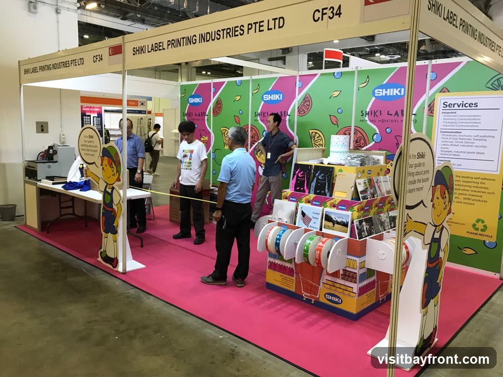 A trade show booth for Shiki Label Printing Industries Pte Ltd. The booth features colorful backdrops with fruit designs and the company logo. Several people are present, including staff and visitors. Displayed items include rolls of labels, printed materials, and a printing machine. A cartoon mascot named Shiki is also featured. A sign lists the company's services.