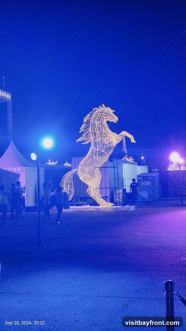 A large, illuminated sculpture of a horse rearing on its hind legs stands in an outdoor area at night. The sculpture is made of white lights. In the background, there are tents, some people, and more lights. The scene is bathed in blue and purple light. A sign with the word 'ENTREE' is visible in the upper left.