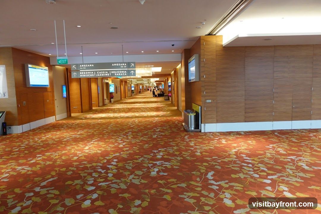 A long, wide hallway with an orange carpet featuring a floral pattern. The walls are paneled with wood-like material. Overhead signs indicate directions to various areas like 'ANGSANA' and 'EXIT'. Digital screens are mounted on the walls.