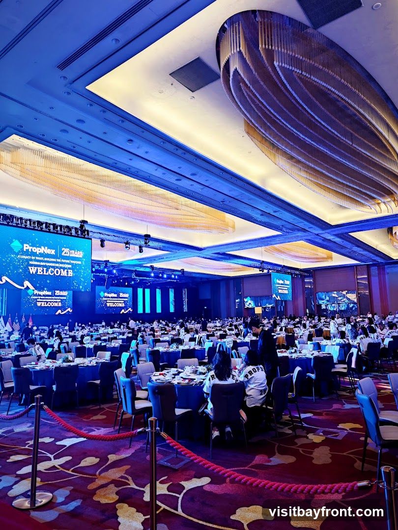 A large ballroom is set up for a gala event with round tables draped in blue tablecloths and chairs around them. A large screen at the front displays "PropNex 25 Years" and "WELCOME." The room is decorated with elaborate chandeliers and illuminated with blue and gold lighting. Attendees are seated at the tables, and some are wearing white shirts with the word "PROP" visible. Red ropes on stanchions guide pathways through the room, which has a patterned carpet.
