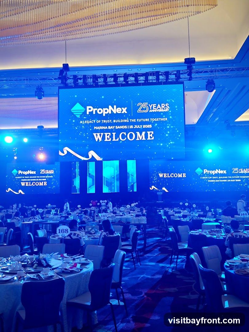 A large screen displays "PropNex 25 YEARS" and "WELCOME" for an event at Marina Bay Sands on July 15, 2025. The room is set up with round tables, chairs, and place settings, indicating a formal gathering or banquet. Several screens are visible throughout the venue, all displaying the same "PropNex" branding and welcome message. Attendees are seated at the tables, and some are wearing shirts with "PropNex" printed on them. The overall ambiance is illuminated by blue lighting.