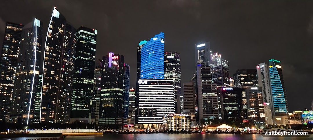 A nighttime view of a modern city skyline with numerous illuminated skyscrapers reflecting in the water below. The buildings display a variety of architectural styles and lighting, with some featuring vibrant blue and purple accents. The scene captures the bustling energy of an urban environment after dark.