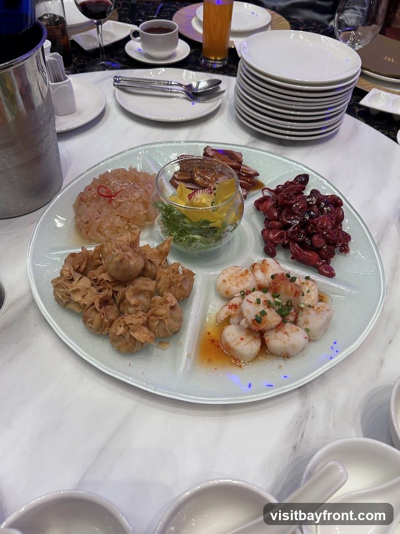 A white platter divided into five sections, each containing a different appetizer. The appetizers include fried dumplings, scallops in sauce, small red octopuses, a salad with edible flowers, and a pinkish shredded salad. The platter is set on a marble-patterned table, with drinks and stacked plates in the background.