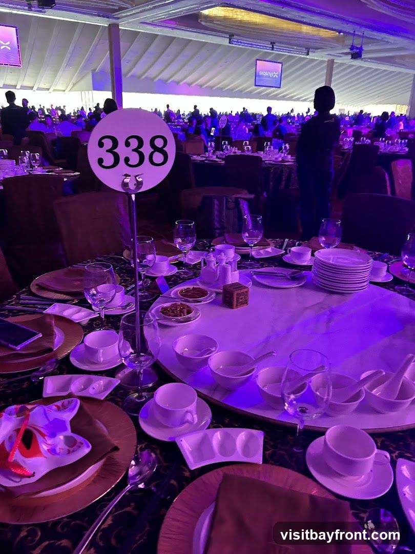 A round banquet table is set for a formal event, with place settings for multiple guests. A large circular sign with the number '338' stands prominently in the foreground. The table is adorned with plates, silverware, glassware, and small bowls, suggesting a meal is about to be served. The room is dimly lit and illuminated by purple and white lights, with a large crowd of people visible in the background, seated at similar tables. Screens displaying logos are visible in the distance.