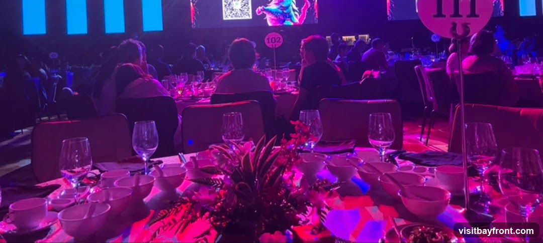 A dimly lit event space with round tables set for a formal dinner. The tables are adorned with white tablecloths, place settings including bowls and wine glasses, and floral centerpieces. Guests are seated at tables in the background, facing a large screen displaying colorful graphics. Table numbers, such as '102' and '111', are visible on stands. The ambiance is festive and elegant, with purple and pink lighting illuminating the scene.