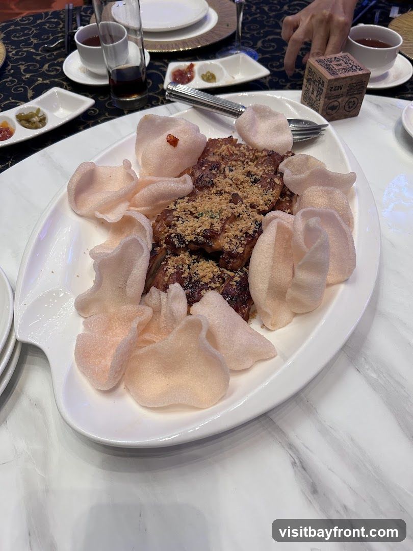 A white oval platter holds a serving of what appears to be fried chicken pieces topped with crushed nuts. The dish is garnished with light pink prawn crackers arranged around the chicken. Small dishes with condiments, a glass of dark liquid, and a cup of tea are visible on the table, along with a small cube-shaped item with text and a logo.