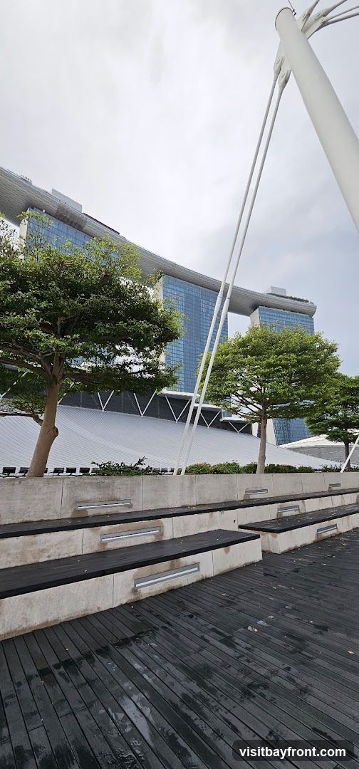 A view of the Marina Bay Sands hotel in Singapore, with its distinctive three towers connected by a sky park. In the foreground, there are tiered wooden benches and trees, suggesting an outdoor seating or viewing area. The sky is overcast.