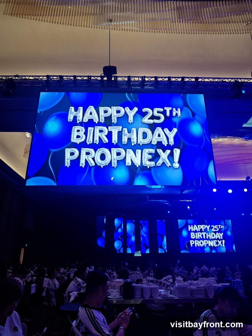 A large screen displays "HAPPY 25TH BIRTHDAY PROPNEX!" with a background of blue balloons. Below the screen, a large audience is seated at tables in a dimly lit venue, with some individuals wearing matching shirts.