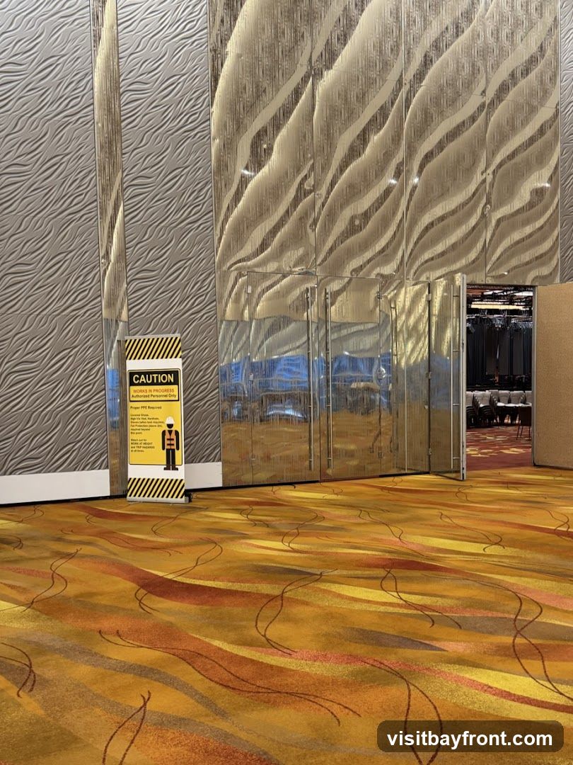 A yellow and black caution sign stands on a patterned carpet in a hallway. The sign reads "CAUTION WORKS IN PROGRESS AUTHORIZED PERSONNEL ONLY" and depicts a person in safety gear. The hallway features textured walls and reflective panels. An open doorway reveals chairs and tables in a room beyond.