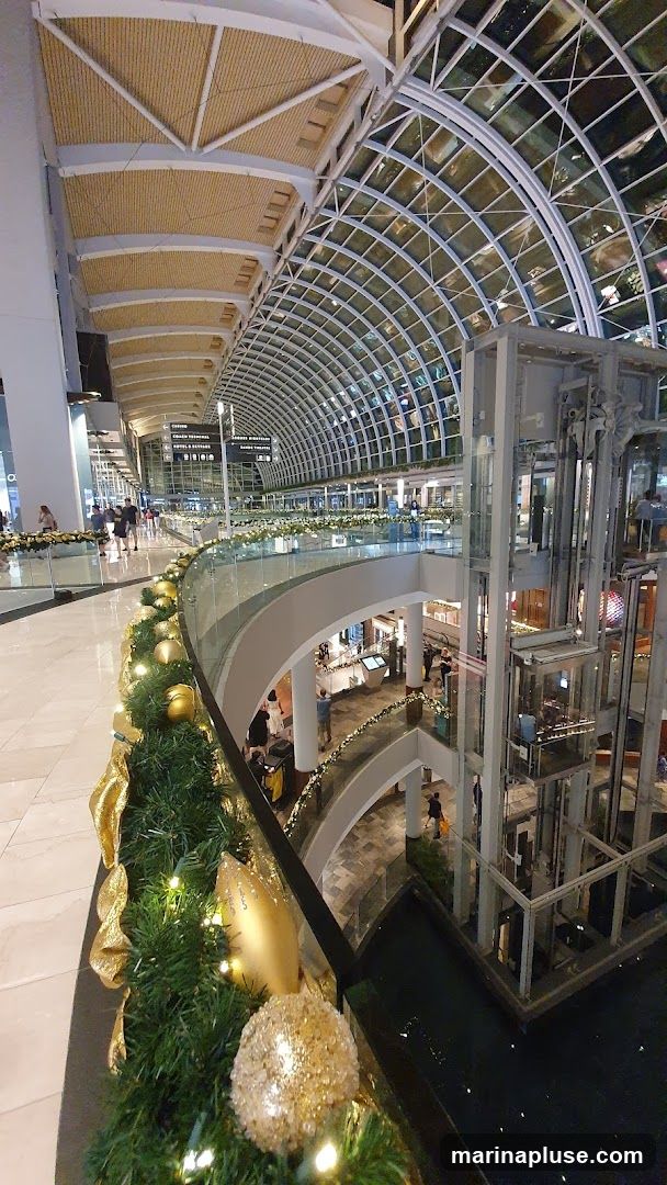An interior view of a multi-level shopping mall with a distinctive arched glass ceiling. The image shows escalators, elevators, and various shops on different floors. Decorative Christmas garlands with gold ornaments and lights adorn the railings. People are seen walking and browsing.