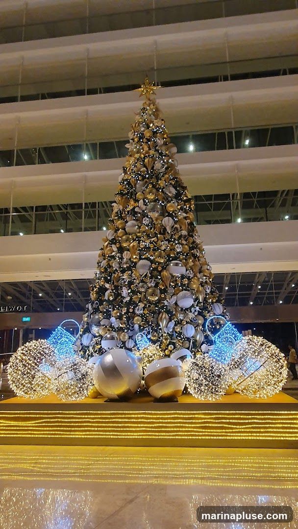 A large, elaborately decorated Christmas tree stands in a well-lit indoor space. The tree is adorned with numerous ornaments in gold, silver, and white, along with strings of lights. At its base, large spherical decorations and illuminated wire sculptures resembling baskets or globes add to the festive display. The background shows multiple levels of a building, suggesting a hotel lobby or a shopping mall.