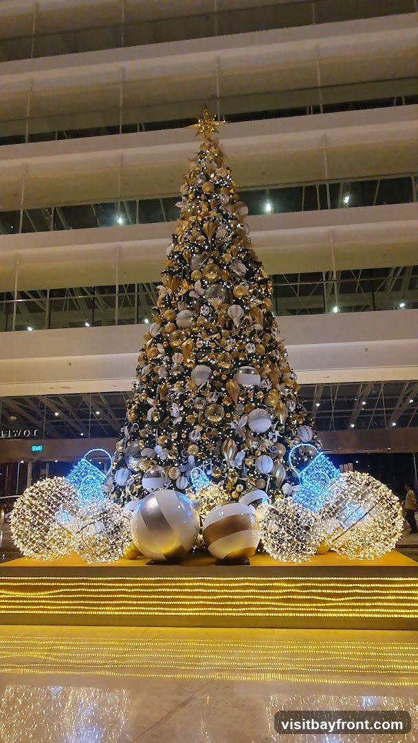 A large, elaborately decorated Christmas tree stands in a well-lit indoor space. The tree is adorned with numerous ornaments in gold, silver, and white, along with strings of lights. At its base, large spherical decorations and illuminated wire sculptures resembling baskets or globes add to the festive display. The background shows multiple levels of a building, suggesting a hotel lobby or a shopping mall.