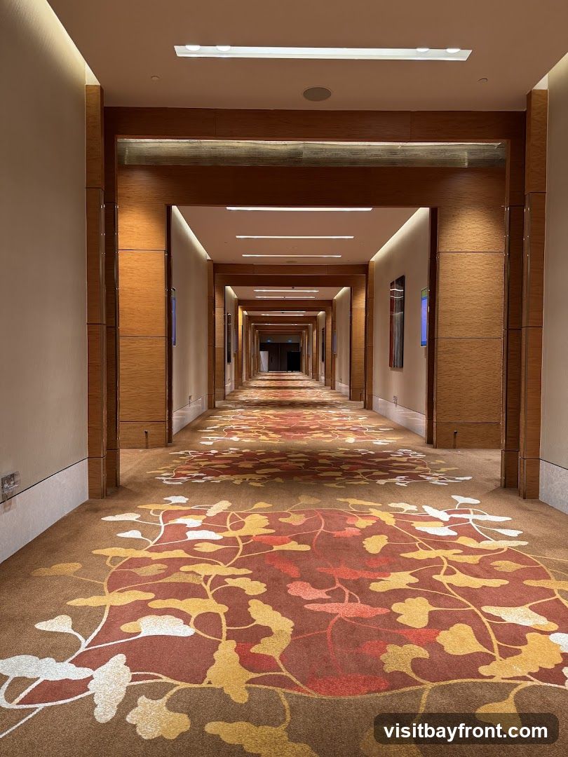 A long, well-lit hotel corridor with a patterned carpet and wooden accents. The carpet features a repeating floral or leaf motif in shades of brown, red, and yellow. The walls are a neutral color, and the corridor is framed by wooden panels and recessed lighting. Several doors are visible along the sides, and artwork hangs on the walls. The perspective draws the eye down the length of the hallway to a dimly lit opening at the far end.