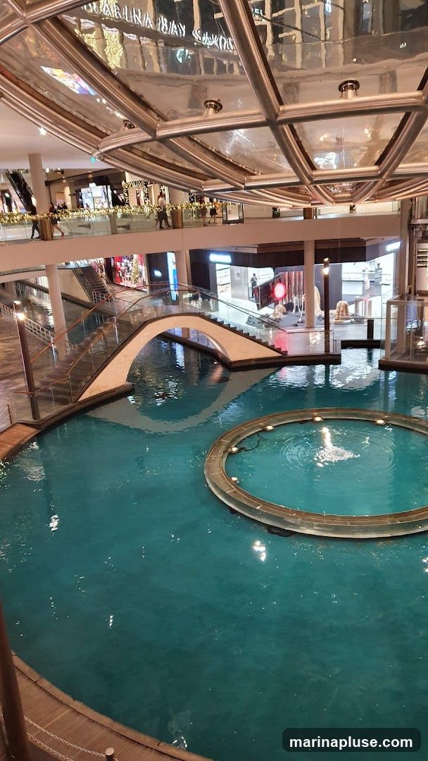 An interior view of a large, multi-level shopping mall. A prominent water feature with a circular fountain is in the foreground, with a white arched bridge spanning over it. People are visible on the upper levels, walking along walkways and near shop entrances. The ceiling has a distinctive geometric design with reflective surfaces. The reflection on the ceiling shows text that appears to read "MARINA BAY SANDS" and other store names.