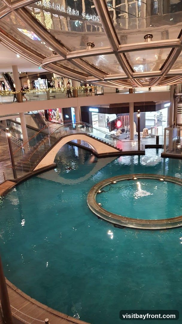 An interior view of a large, multi-level shopping mall. A prominent water feature with a circular fountain is in the foreground, with a white arched bridge spanning over it. People are visible on the upper levels, walking along walkways and near shop entrances. The ceiling has a distinctive geometric design with reflective surfaces. The reflection on the ceiling shows text that appears to read "MARINA BAY SANDS" and other store names.
