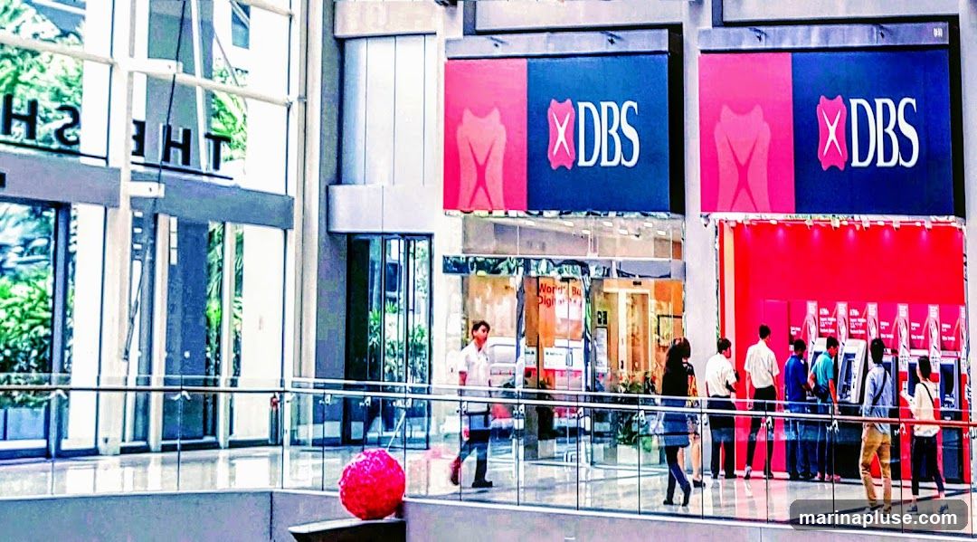 An interior view of a modern bank lobby with multiple DBS branded ATMs lined up against a bright red wall. Several people are seen interacting with the ATMs. Large digital screens displaying the DBS logo are visible above the ATM area. To the left, a glass-enclosed entrance with the partial text "THE SH" is visible. A large, bright pink spherical decoration is placed on a ledge in the foreground.