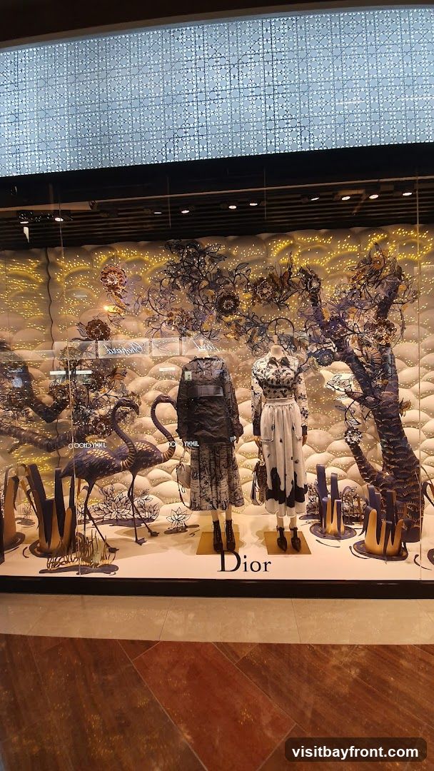 A store window display featuring two mannequins dressed in Dior clothing. The display has a whimsical theme with decorative trees, flamingos, and abstract floral elements. The Dior logo is visible on the display base.
