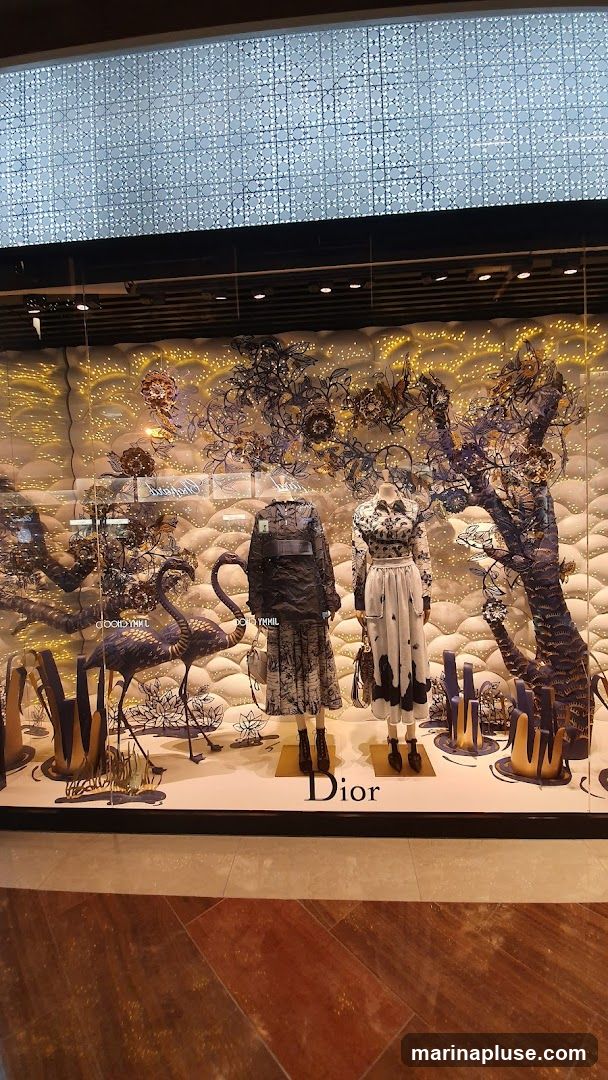 A store window display featuring two mannequins dressed in Dior clothing. The display has a whimsical theme with decorative trees, flamingos, and abstract floral elements. The Dior logo is visible on the display base.