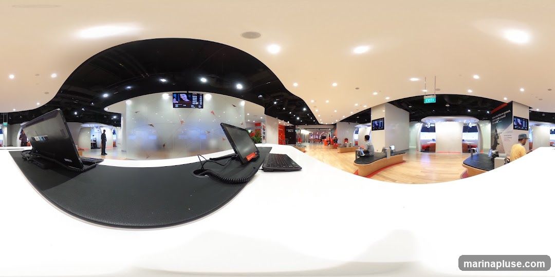 A panoramic view of a bright, modern retail interior. In the foreground, a white counter holds a laptop and keyboard. The background features several curved, illuminated display pods labeled with numbers, suggesting interactive exhibits or service areas. People are seated on benches and standing, interacting with displays or waiting. The space has a clean, minimalist design with wood flooring and white and black accents. An "EXIT" sign is visible overhead.