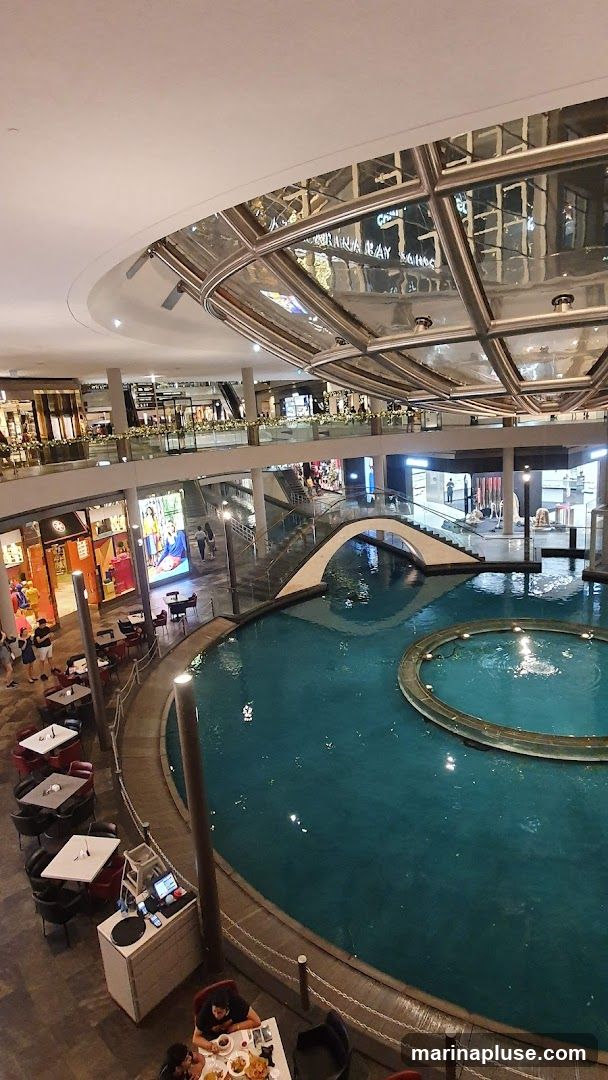 An aerial view of a modern shopping mall featuring an indoor canal with a small arched bridge. Several levels of shops and escalators are visible, along with seating areas and a fountain in the canal.