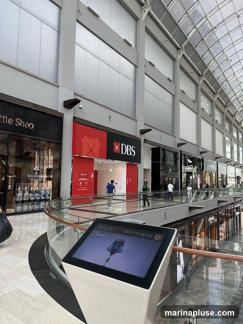An interactive touch screen kiosk is positioned in the foreground of a busy shopping mall. The kiosk displays a Baccarat advertisement with the text "TOUCH TO BEGIN". In the background, various shops are visible, including a DBS bank branch with its prominent red and white logo. Other storefronts and shoppers can be seen on multiple levels of the mall, which features a modern architectural design with a glass ceiling.